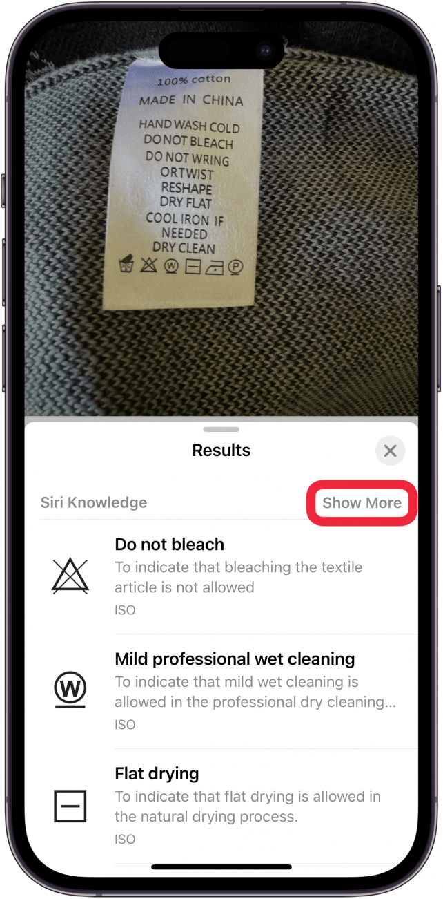 iphone photos app displaying a photo a laundry tag on a sweater, with the visual look up menu open, displaying different laundry symbols and their meaning. there is a red box around the show more button